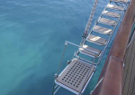 Row of metal seat swings hanging from a ship railing over turquoise water, along a wooden deck edge.