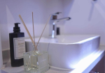 Reed diffuser in a square glass bottle on a bathroom counter beside a dark soap dispenser and a white sink in the background.