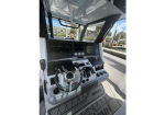 Boat helm with a steering wheel, dual display screens, throttles, and control levers inside a cockpit, view of marina outside.