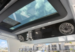 Interior of a white vehicle/boat with a large panoramic sunroof, two circular silver speakers mounted on the ceiling, and blue sky visible outside.