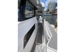Side deck of a white motorboat docked at a marina; gray non-slip steps, railing, with water and city buildings in the background.