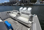 Deck of a speedboat at a marina featuring two white-and-gray cushioned bench seats with cupholders, a coiled rope, and three white outboard engines in the background.