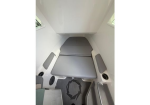 Compact camper van interior with a gray cushioned bed folded into a sleeping area along the wall, flanked by small windows and seating.