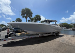 Large white powerboat on a dual-axle trailer parked at a marina on a sunny day, palm trees in the background and vehicles nearby