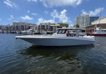 White motorboat cruising in a marina, with docks and modern buildings in the background and two people aboard.