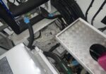 Interior view of a mechanical cabinet showing thick black hoses, blue-labeled clamps, and a circular metal component amid wiring and pipes. It looks like a compact industrial machine under maintenance.