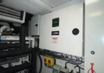 Industrial electrical control cabinet with a green display panel and warning labels in a mechanical room, surrounded by cables and pipes.
