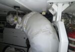 Insulated white industrial duct with protective cover in a mechanical room near a fan and equipment.