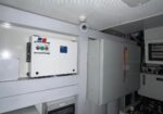 Surveillance camera mounted to the ceiling above an electrical control panel in a utility room, monitoring industrial equipment.