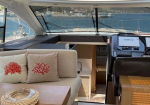 Cozy yacht cockpit lounge with a beige sofa and red coral-pattern cushions, a wooden coffee table, and a helm area to the right with water outside.