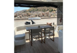 Yacht deck dining area with a wooden table, two director chairs, and a curved white bench; rocky shore and hills in the background.