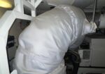 Maintenance worker in white protective suit bending over a machine in a dim industrial engine room.
