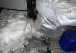 White industrial engine block with silver insulated ducting and a blue jerry can in a metal deck area, likely a mechanical room.