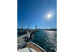 Sunlit marina with sailboats docked along a wooden pier and calm blue water, under a bright sky.