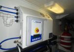 Boat/ship engine-room watermaker unit with blue hoses, control panel, and emergency stop button; a red fire extinguisher is nearby.