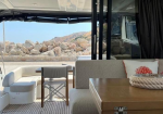 Yacht deck seating area with a wooden table, beige sofa, white cushions with red coral designs, and a rocky coast in the background.