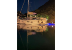 Night scene of a white sailboat docked at a marina, with its reflection shimmering on calm water and a dark hillside in the background.
