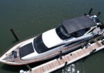 Aerial view of a white motor yacht docked at a wooden pier with a dark canopy over the flybridge and calm water surrounding.