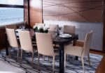 Eight beige upholstered dining chairs around a dark glass table in a modern yacht dining area, with a white floral centerpiece.