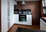 Modern kitchen with white glossy cabinets, dark countertops, and a built-in oven under a stovetop hood, plus wooden wall panels.