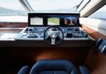 Helm station of a luxury yacht with three navigation screens, steering wheel, and control panels overlooking the sea and mountains in the distance.