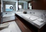 Modern bathroom with a double vanity, marble countertop, and dark wooden cabinets beneath two sinks, mirrors above, and soft lighting. Ocean-view through glass shower enclosure in background.