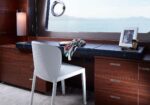 Wooden vanity desk on a yacht, a white chair, scarf draped across the surface, framed photo, and small makeup items, with a sea view through a round porthole window.