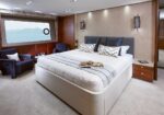 Luxurious bedroom on a yacht with a large white bed, gray headboard, and navy chairs by a window overlooking water.