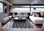 Luxurious yacht lounge with white sectional sofas, a glass coffee table, and a patterned rug, overlooking the ocean.