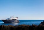 Luxurious black yacht anchored near a rocky shoreline on a calm blue sea, with people on the upper deck under a clear sky.