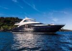 Side view of a large dark blue luxury motor yacht on calm blue water with a rocky coastline in the background.