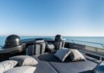 Rear deck of a luxury motorboat with a sunbed and multiple gray cushions, facing the open sea under a blue sky.