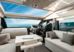 Luxurious yacht salon with beige sofas, a white table, and a helm area overlooking the ocean through large windows and a skylight view of blue sky.