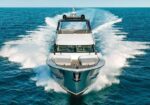 Bow-front view of a white and teal luxury motor yacht speeding through blue ocean with a frothy wake on both sides and trail behind it