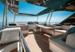 Luxurious yacht deck with beige cushioned seating around a wooden table, ocean view ahead on a sunny day, teak decking and chrome rails visible.