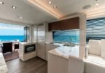 Open-plan galley and lounge on a luxury yacht, with white cabinetry, marble countertops, and an ocean view through large windows.