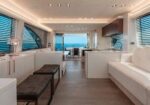 Luxurious yacht living area with white sofas, wooden flooring, and large windows overlooking the ocean; seating and a dining area lead to an outdoor deck.