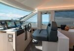 Yacht captain's helm with multiple navigation screens, steering wheel, and a blue-cushioned captain's chair facing the sea window.