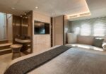 Luxurious bedroom suite with a large bed, gray bedding, and a built‑in media wall facing a seating area by windows with blinds