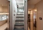 Modern yacht interior with a curved central staircase, flanked by cabins and a seating area on the left.