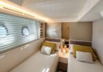 Twin white berths with mustard and white pillows in a modern yacht cabin, porthole windows along the left wall.