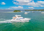 White luxury motor yacht speeds through turquoise water with a small tree-covered island and other boats in the background near a distant city skyline.