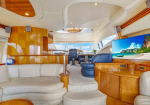 Interior of a luxury yacht with cream leather seating, polished wood paneling, a curved staircase to the helm, and a large TV showing a tropical beach.