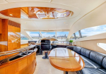 Luxurious yacht salon with polished wood table, curved black leather seating, and the helm visible in the background under skylights.