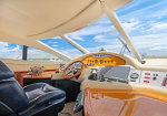 Cockpit of a luxury motor yacht with a wood-grain console, steering wheel, multiple screens, and leather captain chairs under a bright blue sky.