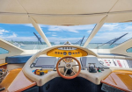 Interior of a luxury yacht cockpit with a wooden steering wheel, beige navigation console, screens, and ocean view through forward windows.