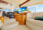 Luxurious yacht salon with cream leather seating, wood cabinetry, and a TV displaying a tropical island; blue sky visible through windows.