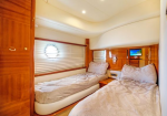 Twin-bed cabin on a yacht with wood paneling, beige bedding, porthole window with blinds, and a small nightstand between beds.