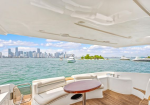 Yacht deck with white seating and a round wooden table, overlooking calm water and a city skyline with several boats.