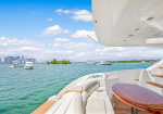 Luxurious yacht deck with white curved seating and a polished wood table, blue water and a distant city skyline on a sunny day.